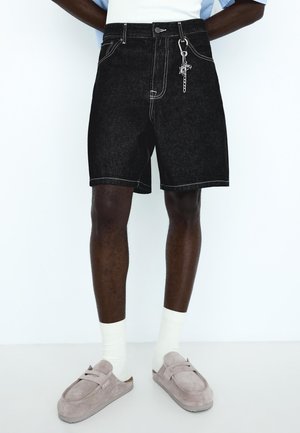 Férfi fekete farmer rövidnadrágot, kulcstartót, fehér zoknit és szürke szarvasbőr slip-on cipőt visel, egyszerű fehér háttér előtt áll.