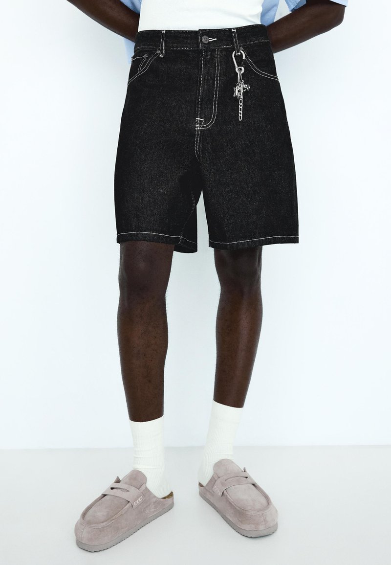 Homme portant un short en denim noir avec un porte-clés, des chaussettes blanches et des chaussures en daim gris, debout devant un fond blanc uni.