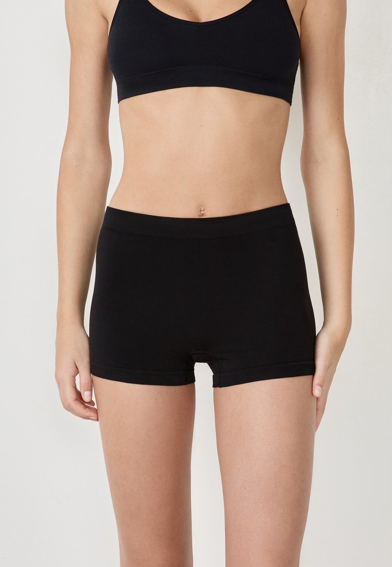 Femme portant un short noir sans couture et un soutien-gorge de sport noir assorti, debout devant un fond clair uni.