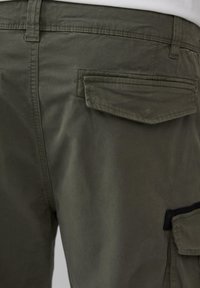 Zielone spodnie cargo w odcieniu oliwkowym charakteryzują się teksturowanym wykończeniem, licznymi kieszeniami oraz przeszyciami z tyłu, podkreślając praktyczny design.