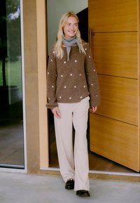 Bruine gebreide trui met witte stippen, gecombineerd met roomkleurige wijde broeken en donkere loafers. De outfit wordt aangevuld met een grijze sjaal.