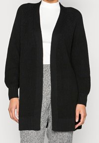 Černý pletený kardigan s otevřeným předním dílem, žebrovanými manžetami a lemem. Texturovaná látka a uvolněný střih, nošený přes bílý top a vzorované kalhoty.