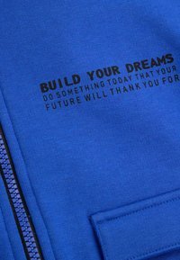 Niebieska bluza z suwakiem. Na przodzie czarny nadruk: "BUDUJ SWOJE MARZENIA" oraz motywujący cytat poniżej.