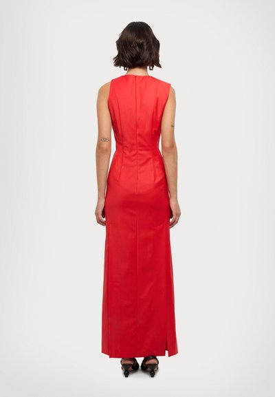 Robe maxi sans manches rouge avec une silhouette ajustée, fermeture éclair à l'arrière, fentes latérales et tissu lisse. Portée avec des chaussures à talons noires.