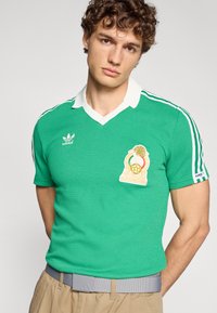 Πράσινο πόλο μπλουζάκι από υφασμάτινη υφή, με λευκό γιακά, λευκές ρίγες στα μανίκια και κεντημένο έμβλημα στο στήθος.