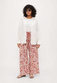 Cămașă albă cu mâneci lungi peste un top alb fără mâneci, asociată cu pantaloni evazați roșii cu un model. Ochelari de soare negri și sandale albe completează ținuta.