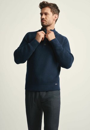Granatowy dziany pulower z wysokim kołnierzem i trzema guzikami. Żebrowana tekstura, długie rękawy i lekko dopasowany fason.