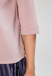Dettaglio della manica di una blusa testurizzata rosa chiaro con polsini cuciti, indossata con una gonna plissè scura su uno sfondo neutro.