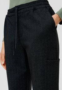 Zwarte geklede broek met een elastische tailleband en een trekkoord. Zichtbare zijzakdetails. Gemaakt van geweven stof met een subtiel patroon.