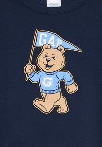Marineblauer Pullover mit einem Cartoon-Bären, der einen hellblauen Pullover mit einem "G" trägt und eine Fahne hält, auf der "GAP" steht.