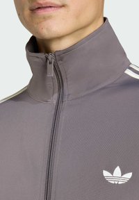 Grå idrottsjacka med hög krage, dragkedja fram och vitt logotyp. Har vita ränder längs axlarna och strukturerad tyg.