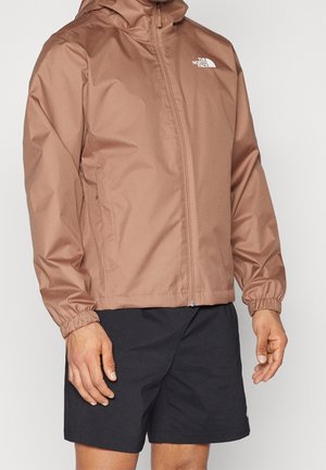 Bruine waterdichte jas met ritssluiting, capuchon, elastische manchetten en logodetail op de borst. Draagt zwarte korte broek.