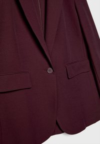 Burgundy blazer dengan tekstur halus, penutupan kancing tunggal, dua saku depan, kerah bergerigi, dan potongan yang disesuaikan.