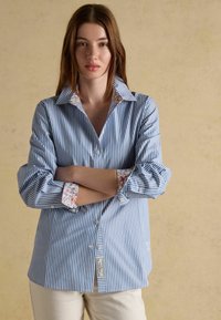 Young woman with long brown hair wearing a blue and white striped button-up shirt with floral inner cuffs, standing with arms crossed.