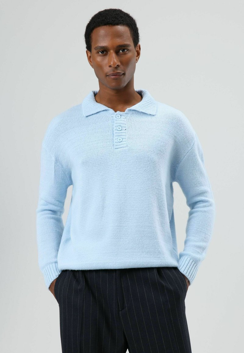 Lichtblauwe gebreide trui met een polokraag, voorzien van een vierknopenpanden en geribde manchetten, gecombineerd met donkere broek met fijn streepje.