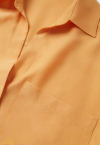 Gros plan d’une chemise orange clair montrant le col, les coutures avant et une poche poitrine sur un tissu lisse.