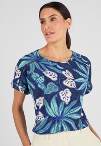 T-shirt azzurro a maniche corte con grandi motivi di foglie tropicali verdi e scollatura rotonda. Tessuto morbido con risvolti sulle maniche e dettagli di cucitura discreti.
