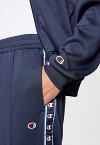 Μπλε ναυτικό τζάκετ φόρμας με φερμουάρ και ελαστικά μανίκια. Εμβληματικό λογότυπο στο μανίκι με πολύχρωμες ρίγες στο πλάι. Λείο ύφασμα.