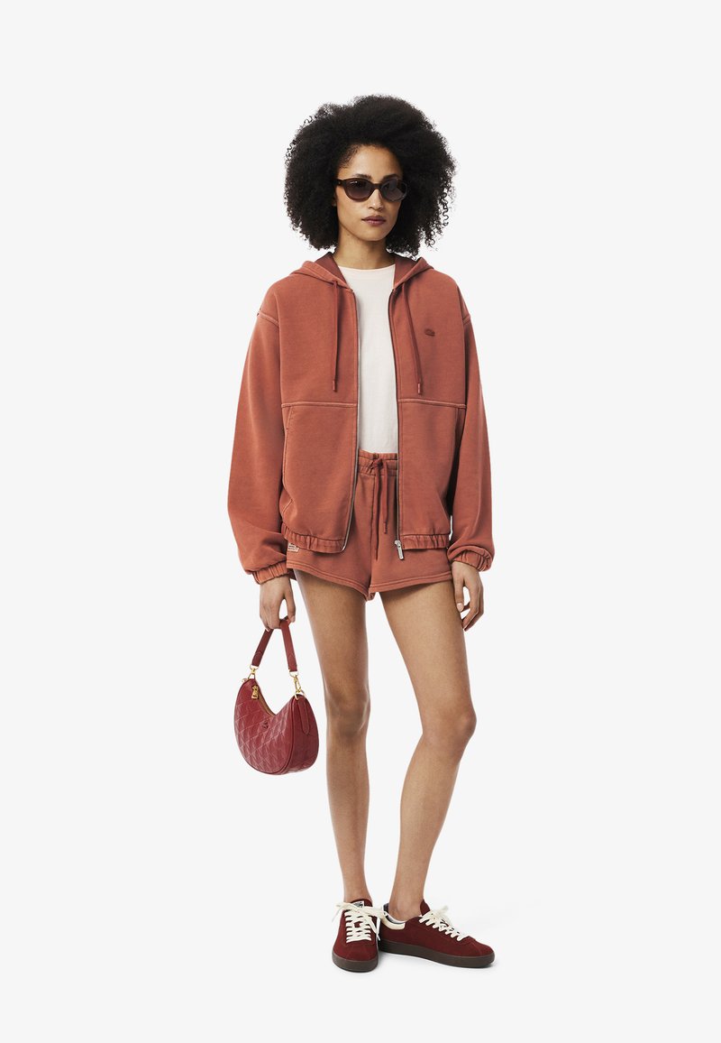 Haut à capuche zippé de couleur rouille avec de grandes poches avant, shorts assortis, sac à main matelassé rouge et baskets bourgogne avec des lacets blancs.