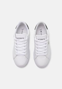 Fehér, fűzős sportcipők sima szintetikus anyagból, fekete díszítéssel a sarkon és "LACOSTE" felirattal az oldalán.