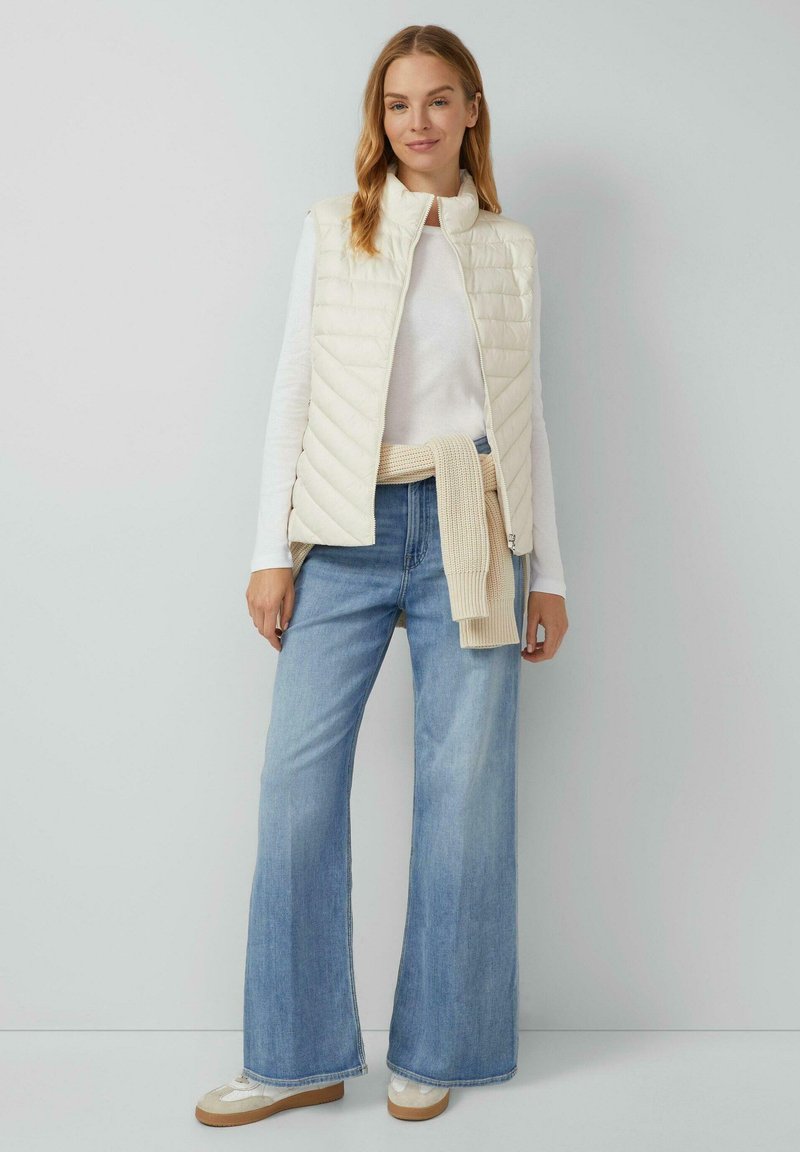 Lichtgewicht crèmekleurige puffer vest met horizontale quilting, gecombineerd met een witte langemouwen top en brede blauwe jeans. Beige gebreide trui gestrikt in de taille.