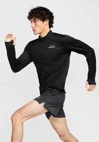 Schwarzes langärmliges Sportoberteil mit Viertelreißverschluss, versehen mit dem Nike-Logo, kombiniert mit schwarzen Shorts, in einer dynamischen Laufpose.