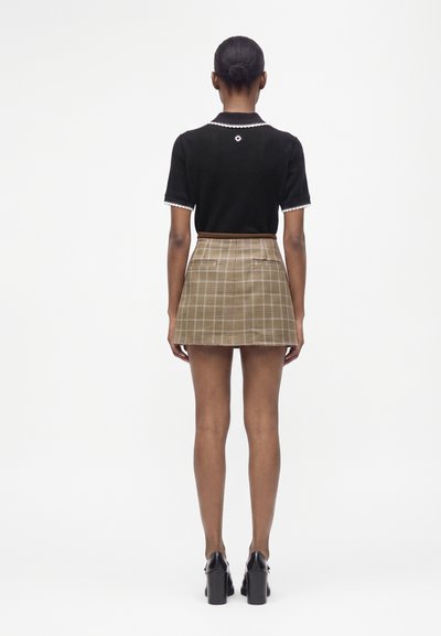 Femme aux cheveux en chignon, portant un haut noir à manches courtes avec une bordure blanche, une jupe marron à carreaux et des chaussures à talons noires, debout de dos.