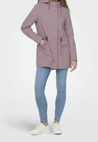 Kobieta w mauve kurtce z kapturem i sznurkami, w jasnoniebieskich dopasowanych dżinsach i białych trampkach, stoi z jedną ręką w kieszeni.