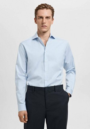 Camisa de vestir azul claro con finas rayas verticales, cuello abotonado y mangas largas, combinada con pantalones oscuros. Textura de tela suave.