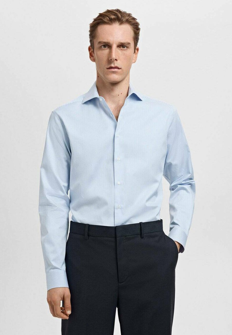 Camicia azzurro chiaro con sottili righe verticali, colletto button-down e maniche lunghe, abbinata a pantaloni scuri. Tessuto dalla consistenza liscia.