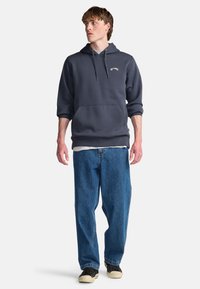 Gray hooded sweatshirt with a kangaroo pocket, paired with loose blue jeans and black sneakers. Features a minimalist logo on the chest.
