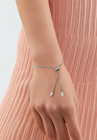 Zilveren kettingarmband met verstelbare lengte, met twee puntige accenten aan de uiteinden. De achtergrond is een gestructureerde roze stof.