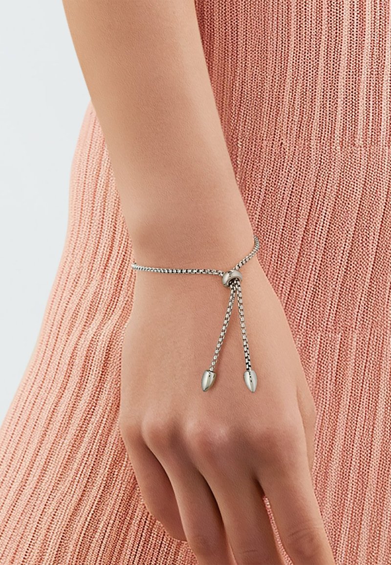Zilveren kettingarmband met verstelbare lengte, met twee puntige accenten aan de uiteinden. De achtergrond is een gestructureerde roze stof.