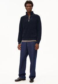 Mann trägt einen dunkelblauen Reißverschluss-Strickpullover, ein graues Hemd darunter, lockere blaue Jeans und schwarze Quasten-Lederschuhe und steht vor einem weißen Hintergrund.