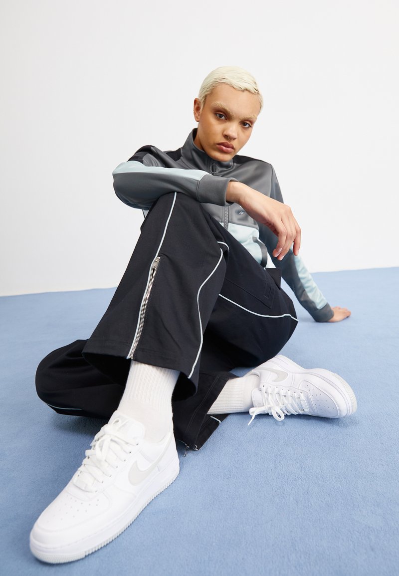 Persona con pelo rubio corto sentada en el suelo azul, vistiendo pantalones negros con ribete blanco, chaqueta gris con cremallera y zapatillas blancas con calcetines.