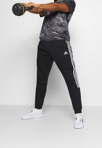 Zwarte joggingbroek met witte zijstrepen en een elastische tailleband, gecombineerd met een grijs camouflage t-shirt en witte sportschoenen.
