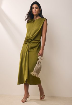 Femeie purtând o rochie midi fără mâneci, verde măsliniu, cu detaliu drapat, ținând o poșetă cu șnur și franjuri auriu și purtând sandale aurii cu toc.