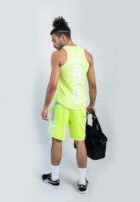 Canottiera sportiva gialla neon con testo bianco e pantaloni corti abbinati. Accenti bianchi sui pantaloni corti. Borsa da palestra nera tenuta in una mano.