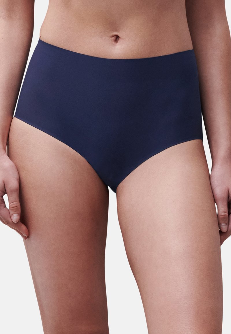 Nainen pukeutuneena korkeavyötäröisiin laivastonsinisiin saumattomiin alusvaatteisiin, esittäen vartalon lantiosta reiden keskivaiheille selkeällä taustalla.