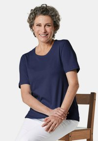 Marineblauw T-shirt met korte mouwen en een ronde hals, gedragen door een persoon die op een houten stoel zit, gecombineerd met witte broek.