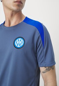 Camiseta deportiva gris con acentos azules, logotipo redondo en el pecho izquierdo, mangas cortas, tejido texturizado y cuello redondo.