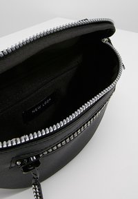 Musta synteettinen käsilaukku, jossa on vetoketjukiinnitys ja hopeanväriset yksityiskohdat. Sisältää vuoratun sisäosan, jossa on pieni vetoketjutasku ja brändilappu.