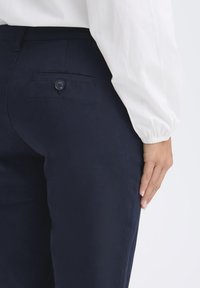 Navyblaue Hose aus leichtem Stoff, mit einer Gesäßtasche und einem Knopf sowie einer glatten Textur. Hand an der Seite abgelegt.