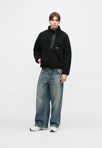 Μαύρο fleece πουλόβερ με μισό φερμουάρ, μπροστινή τσέπη και λείο μαύρο πάνελ; συνδυασμένο με φαρδιά, ξεθωριασμένα μπλε τζιν και λευκά sneakers.