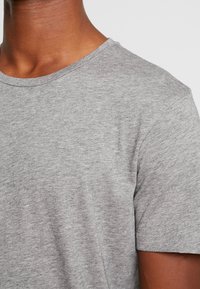 Grå T-shirt med korte ærmer og rund hals, blød tekstur og subtil meleret mønster, der viser et nærbillede af skulderområdet.