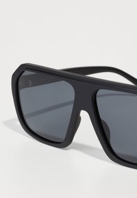 Occhiali da sole neri con finitura opaca, forma rettangolare grande, lenti scure e aste robuste. Design minimalista con linee pulite.
