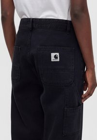 Detailný pohľad na osobu, ktorá nosí čierne nohavice Carhartt s viditeľnými zadnými vreckami a logom na pravom vrecku.