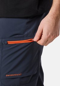 Laivastonsiniset cargoshortsit, joissa on oranssi vetoketjupussi, valmistettu kevyestä kankaasta. Ominaisuuksiin kuuluu "SWEDEMOUNT" brodeerattuna oranssilla pussin lähellä.