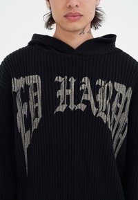 Felpa nera a coste con un grande testo grafico metallico argento sul davanti. Il design include un cappuccio spazioso e un tessuto testurizzato.