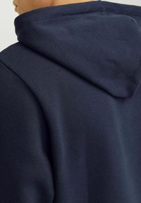 Námornícky modrá mikina s kapucňou, vyrobená z mäkkej zmesi bavlny, s veľkou zaoblenou kapucňou a hladkou štruktúrou s minimálnymi švami a detailami šitia.